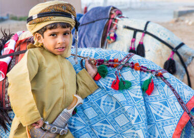 Muscat, Oman - Feb 4, 2017: Young Omani boy dressed in traditional clothing posing next to his camel.